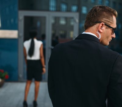 Personal guard in sunglasses and security earpiece, back view, black business woman on background. Bodyguard is a risky profession, protection of VIPs, guarding